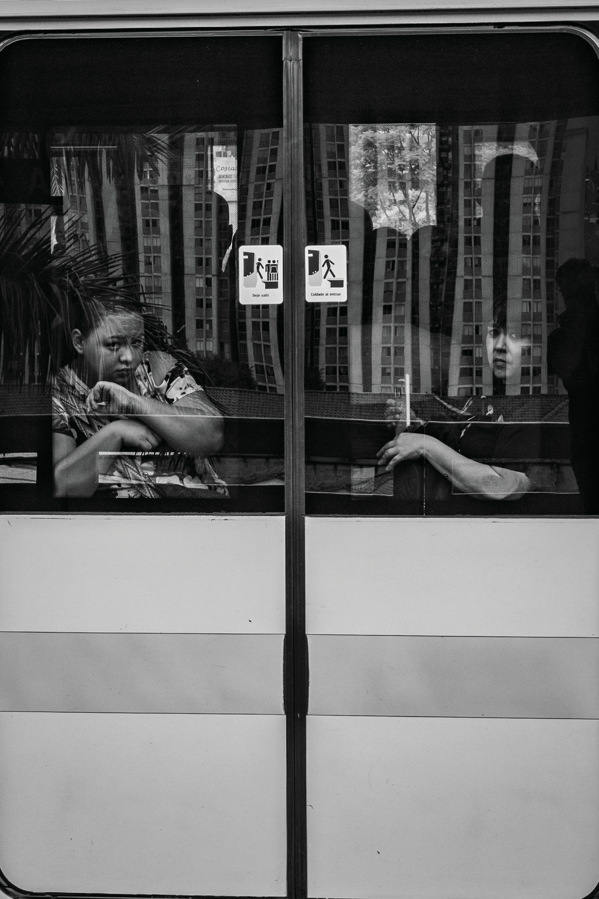 Two passengers seen through the divided windows of a city bus, one glancing toward the camera.