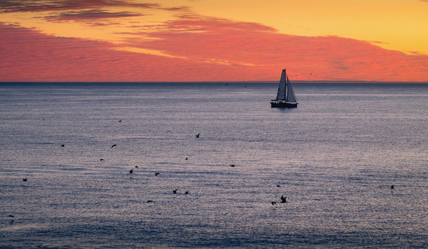 A sailboat and scattered seabirds beneath a vivid orange and coral sunset over the Pacific Ocean.