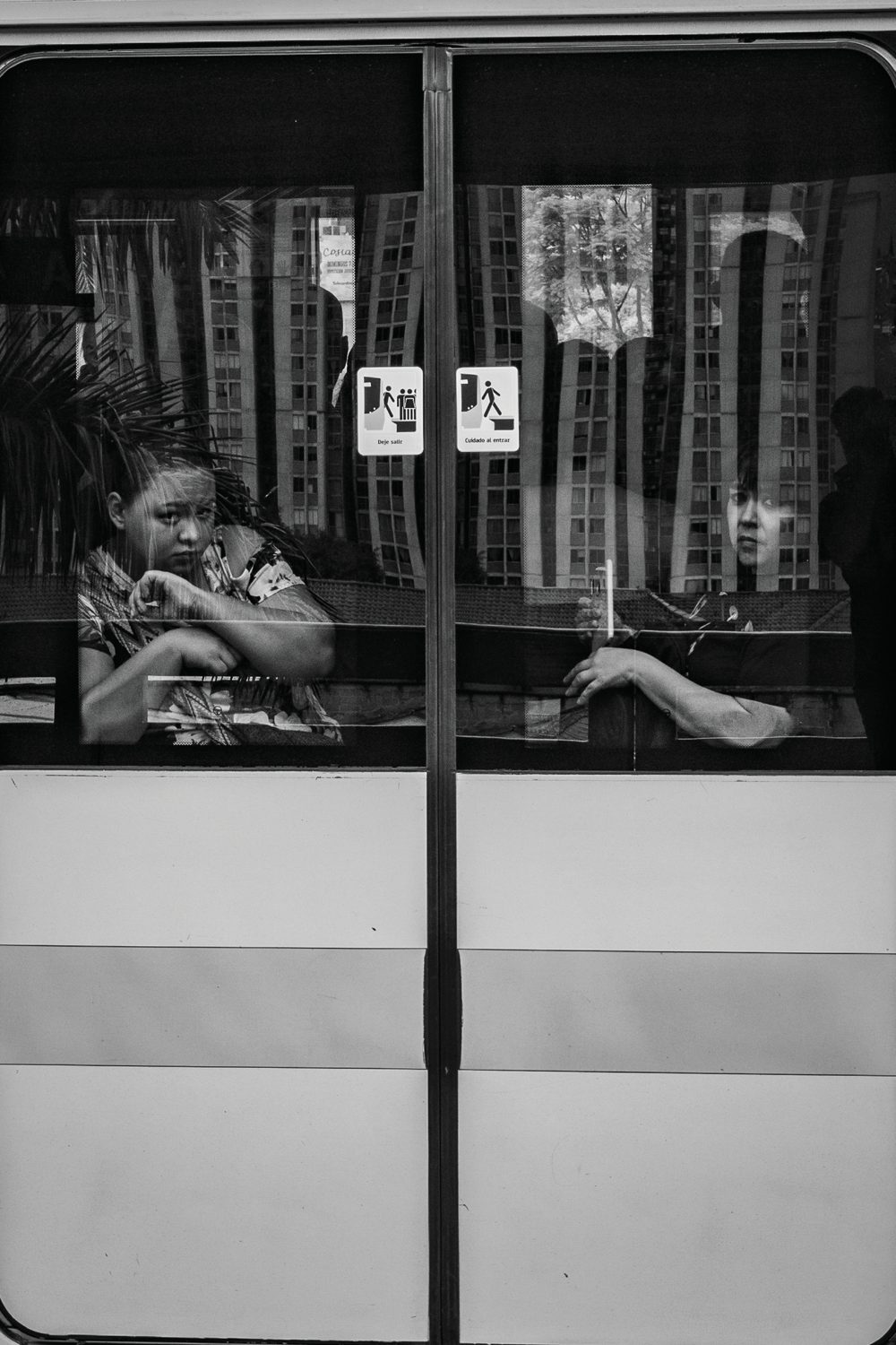 Two passengers seen through the divided windows of a city bus, one glancing toward the camera.