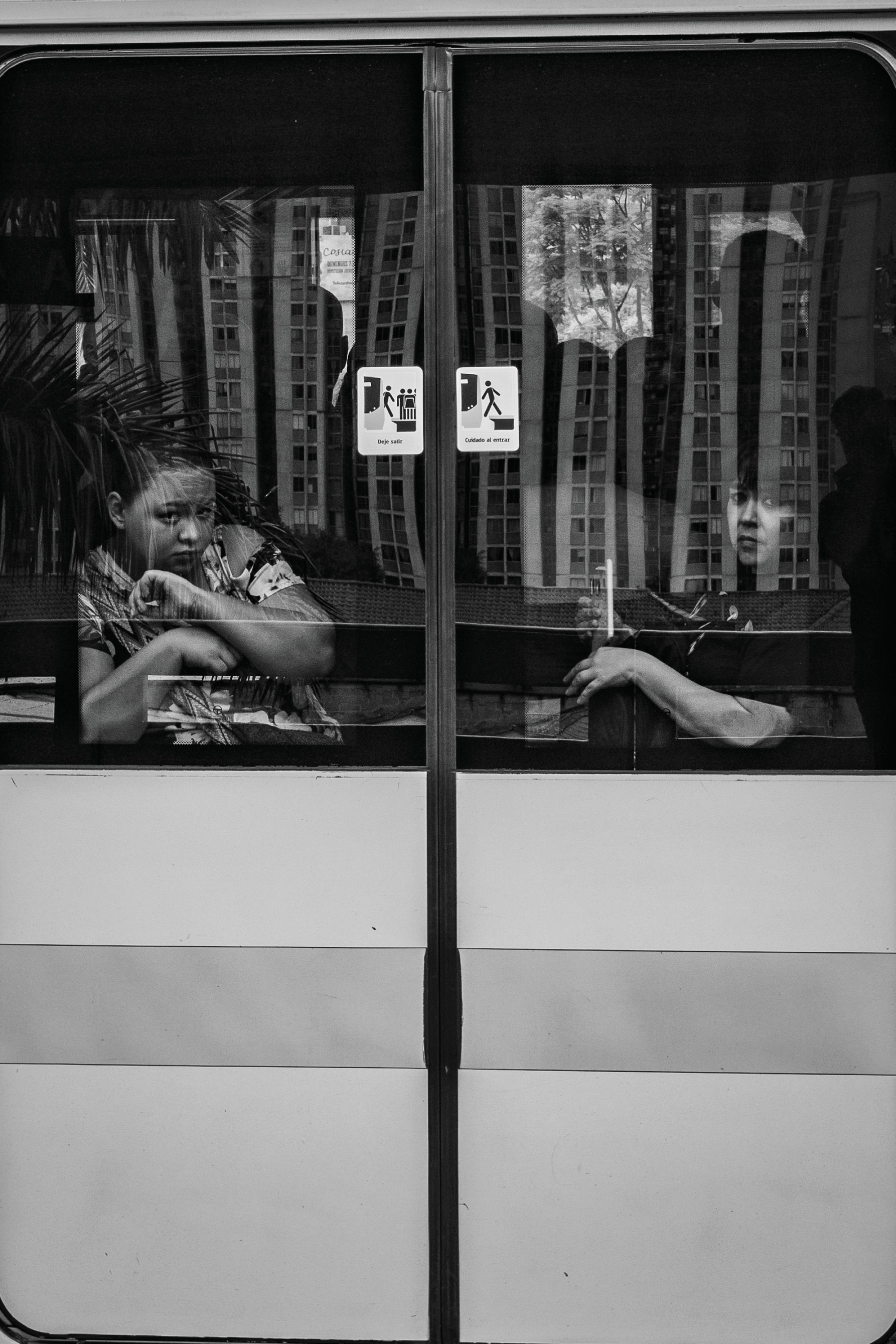 Two passengers seen through the divided windows of a city bus, one glancing toward the camera.
